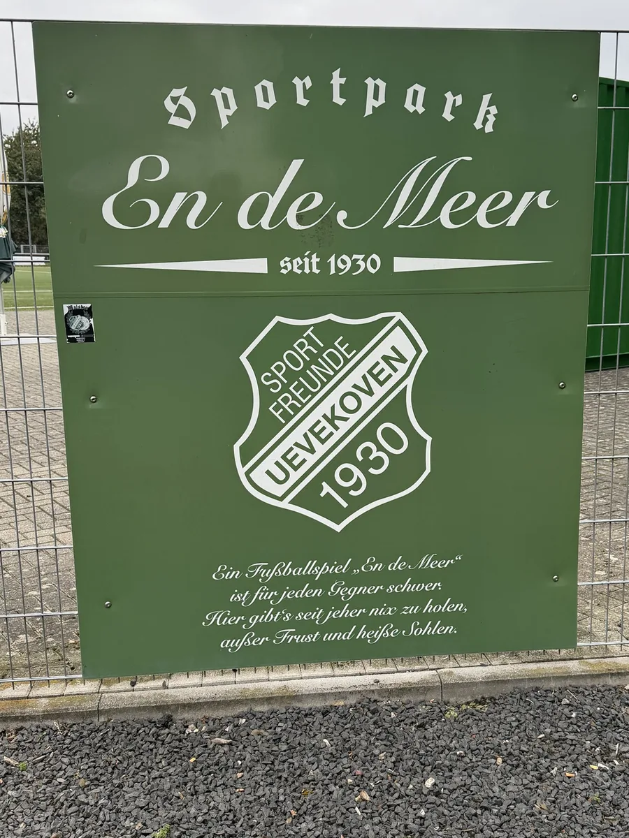Grüne Tafel am Eingangstor zum Kunstrasen: Sportpark En de Meer, seit 1930, mit Vereinswappen und Platzspruch