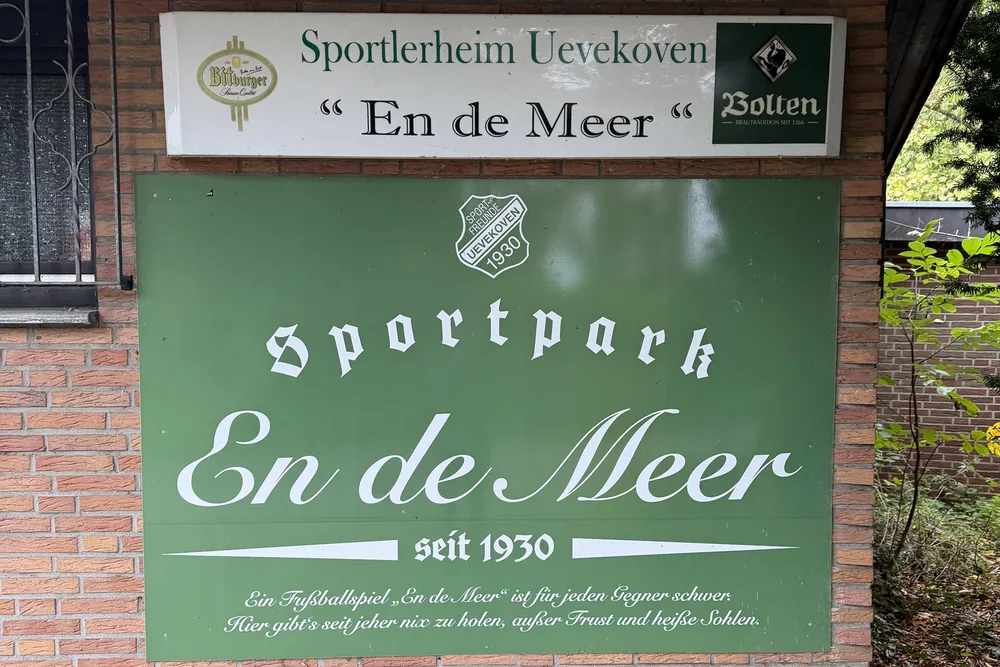 At the passage to the grass pitch: the sign “Sportlerheim Uevekoven, En de Meer” and below it the green board “Sportpark En de Meer, since 1930” on the brick wall